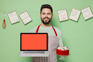 Wall Mural - Young male chef confectioner baker man in striped apron hold cake use work on laptop pc computer with blank screen workspace area isolated on plain pastel light green background. Cooking food concept.