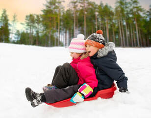 Poster - childhood, sledging and season concept - happy little kids sliding on sled down snow hill in winter over pine forest on background