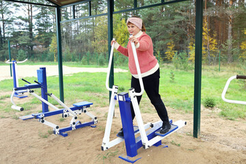 Chubby joyful girl performs sports exercises on street simulators outdoors