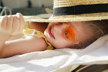 Adorable toddler girl laying on sun bed and hiding from sun under straw hat. Happy summer time childhood concept.