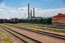Railroad Station. Trains With Open Wagons On Tracks, Factory In Red Bauxite Clay (aluminium Ore) Dust. Three Smoke Stacks Of Power Plant On Background