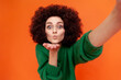 © khosrork - Positve friendly woman with Afro hairstyle in green casual style sweater making selfie POV, sending air kisses to followers while having livestream. Indoor studio shot isolated on orange background.