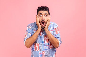 Portrait of astonished bearded man in blue shirt looking with big eyes, holding hands on face and screaming in amazement, shocked by sudden event. Indoor studio shot isolated on pink background.