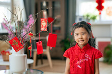 Happy Cute Asian Little Girl In Red Dress Looking At You While Standing In Living-room Decorated For New Year