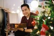 © Mallika - Smiling Asian man in red sweater unboxing gift box at home,Happy Asian man in Christmas Day concept.