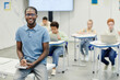 © Seventyfour - Portrait of young African-American teacher sitting on desk and smiling at camera with children in background, copy space