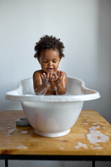 black toddler sitting in bath with foam. bathing without tears. cute emotional baby is engaged in hy