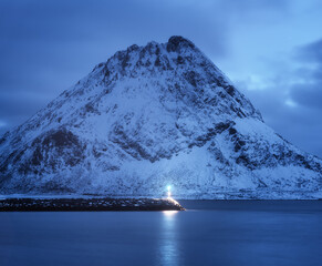 Wall Mural - Lighthouse, sea and beautiful snow covered mountain and cloudy blue sky reflected in water at dusk. Winter landscape with lighthouse, snowy rock at night. Lofoten islands, Norway at twilight. Nature