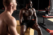 © Roman - Side View On African And Caucasian Males Sitting On Plyobox Having Rest Talking, Laughing, Two Young Strong Shirtless Men In Dark Gym. Sport And Fitness, Cross Fit Concept. Motivation Concept