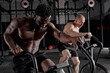 two diverse handsome muscular men doing hard bike training in gym, shirtless strong african american and caucasian motivated sportsmen concentrated on cardio training, workout. side view, indoors