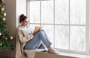 Canvas Print - Beautiful young woman with book near window at home on Christmas eve