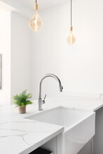 A Kitchen Sink Detail Shot With Glass Pendant Lights Hanging Above The White Granite Countertop, Apron Sink, And Chrome Delta Faucet. 