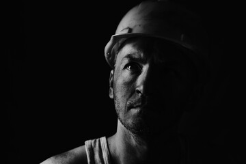 Dirty coal miner in a yellow hard hat on a dark background in a black and white photo.