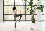 Fototapeta Londyn - Young multiracial woman kicking leg up doing online fitness training at home living room. Copy space