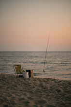 Sitting And Fishing Pole On Coastline During Sunset. No Man Fishers Place On The Seaside At The Summer Evening