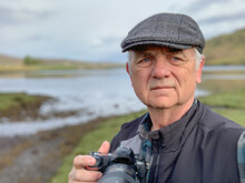 Portrait Of A Man Wearing A Flat Cap Standing In Rural Landscape Taking Photos, Isle Of Skye, Inner Hebrides, Scotland, UK
