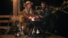 Beautiful People Warm Their Hands By The Fire.  Beautiful Woman In A Hat And A Man With A Beard Are Sitting On The Terrace And Warming Their Hands At The Barbecue