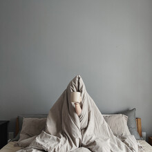 Woman Sitting In Bed Under Blanket, Holding Mug With Coffee In Hand. Happy Morning Breakfast. Freelancer, Pandemic Quarantine Day Lifestyle Concept. Minimalist Bedroom Interior Design