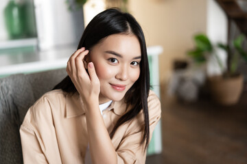 Portrait of asian woman smiling, sitting at home. GIrl touches her hair and looks cute at camera