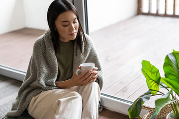 Wall Mural - Young sad asian woman sitting on floor near balcony and looking at tea mug thoughtful