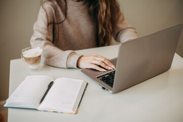 Young brunette woman with long hair uses a laptop at home. There is a mug of coffee and a notebook with a pen on the white table. No face visible