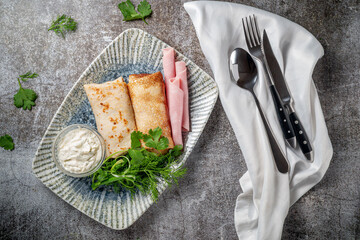 Poster - Pancakes filled with ham in a plate with parsley and dill greens and sour cream on a gray stone table