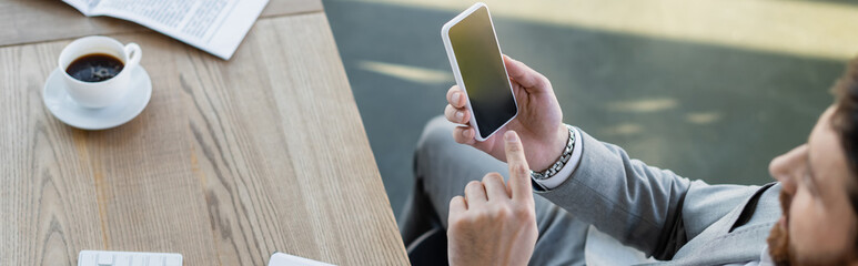 High angle view of blurred businessman pointing at smartphone near newspaper and coffee in office, banner.