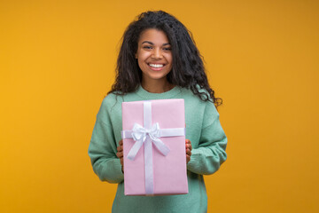 Studio portrait of beautiful smiling african american girl posing with decorated gift box in hands, isolated over bright colored yellow background