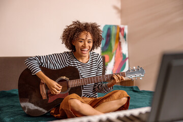 Joyful female musician sitting on bed and laughing while playing melody on acoustic musical instrument