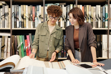 Wall Mural - Young confident brunette female shop assistant helping client to choose sample of new wallpapers in contemporary studio of interior design