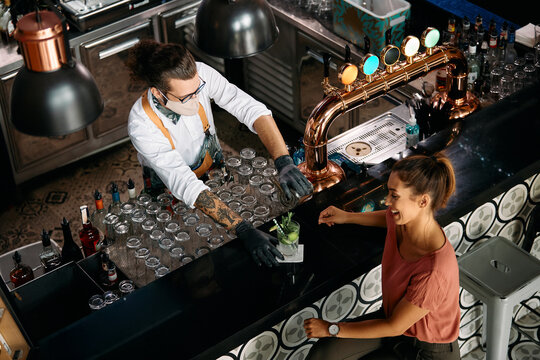 Above view of barman serving cocktail to female customer at bar counter while and wearing face mask due to COVID-19 pandemic.