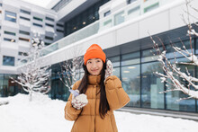 Young Beautiful Asian Woman Listens To Music From Headphones While Walking On A Winter Snowy Day, Uses An Online Application For The Phone