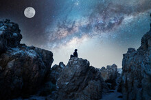 Person On The Rock Outdoors Meditating Or Praying At Night Under The Milky Way And Moon	