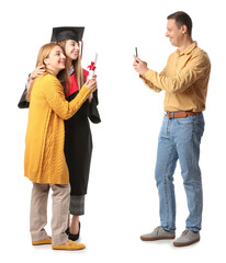 Wall Mural - Happy man taking photo of his wife and daughter in graduation gown and with diploma on white background