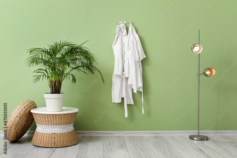 Houseplant with table, lamp and white bathrobes hanging on green wall