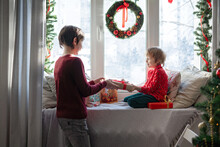 Two Brothers With Gift Boxes Watching The Snow Fall Through A Window In Festively Decorated Room. Waiting For Santa
