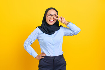 Wall Mural - Portrait of smiling young Asian woman showing peace gesture and looking at camera isolated on yellow background