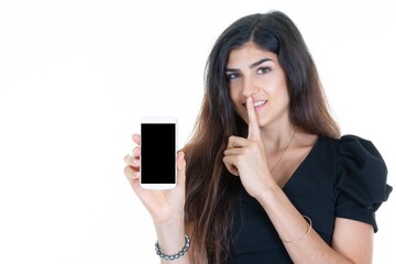 Wall Mural - beautiful woman makes secret silence gesture on lips while holding smartphone which has empty black screen of phone on white background