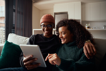 Adult couple, multi-ethnic, smiling while using tabe on sofa in modern apartment