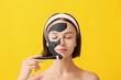 Young woman applying activated charcoal mask on her face against color background