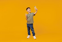 Full Body Little Small Happy Boy 6-7 Years Old In Green T-shirt Do Selfie Shot On Mobile Cell Phone Show V-sign Gesture Isolated On Plain Yellow Background. Mother's Day Love Family Lifestyle Concept.