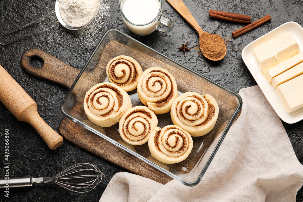 Baking dish with uncooked cinnamon rolls and ingredients on black background