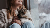 Young cheerful girl sitting at home on the windowsill in a warm sweater playing with a gray cat on a winter day