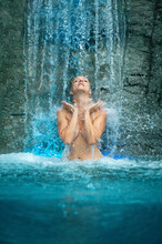 Beautiful Young Cute Sexy Blonde Woman Under The Splashing Water Of The Waterfall In The Spa Wellness Pool Enjoys The Falling Water, Lets The Rippling Water Patter On Her Raised Hands