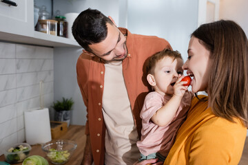 Cheerful man standing near son feeding mother with cherry tomato in kitchen