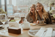 Close-up Portrait Of Two Female Friends In Strict Suits Laughing Drinking Coffee And Wine On Terrace Outside At Summer Street Cafe On Background Buildings Old Tbilisi City, Georgia. High Quality Photo