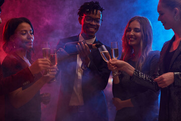 Wall Mural - Group of cheerful young people pouring champagne in smoky nightclub while enjoying party