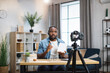 © sofiko14 - African man in headset sitting at desk with book in hands and using modern laptop with digital camera for recording online lessons. Concept of people, technology and distance learning.