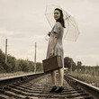 A young woman with an umbrella and a suitcase walks away along the rails of the railway and looks back at goodbye.