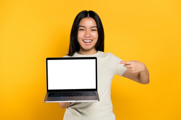 Excited chinese positive young woman in casual t-shirt holding open laptop with blank white screen and points a finger at him, stands on isolated orange background, looks at camera, mockup, copy space